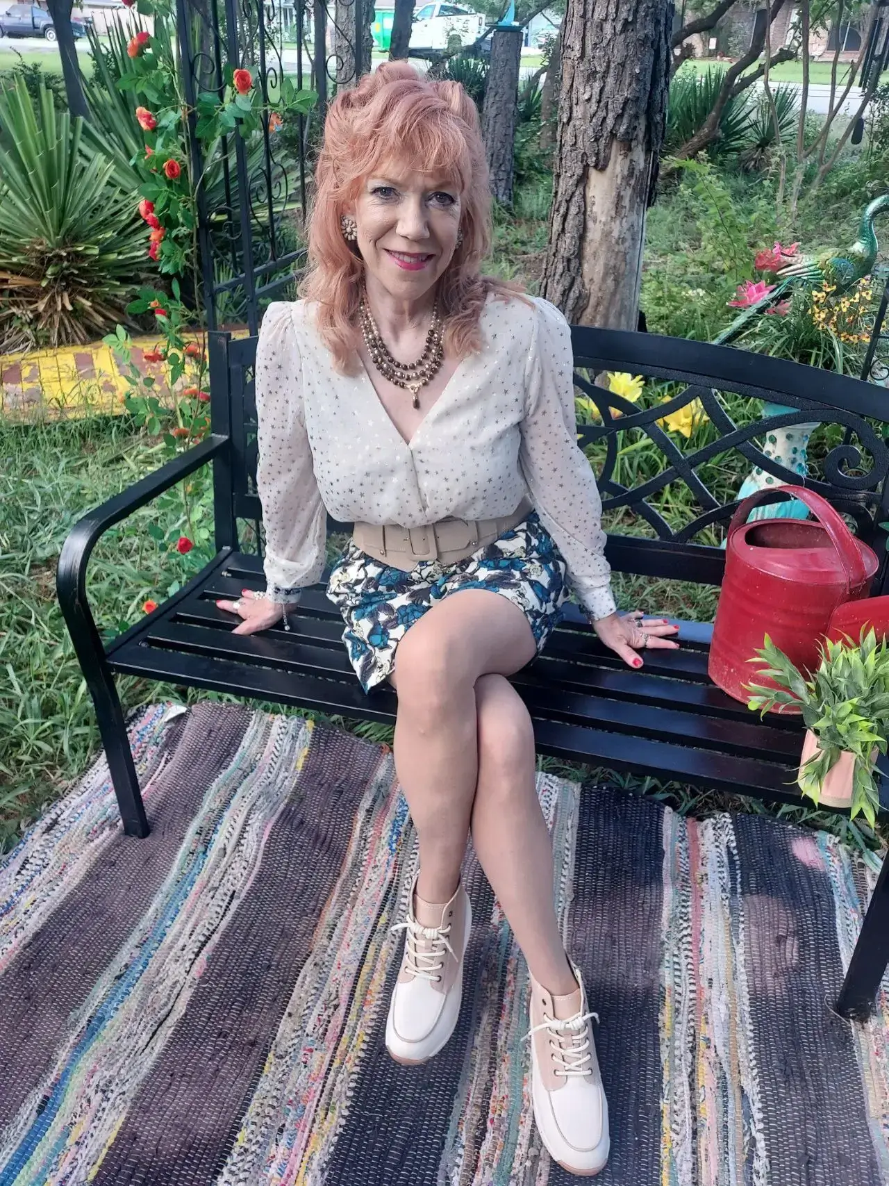 A woman sitting on a garden bench surrounded by plants and a watering can.